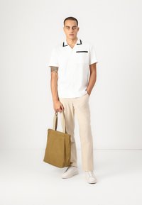 White short-sleeve shirt with black accents, beige pants, and white sneakers. Holding a brown canvas tote bag with soft texture.
