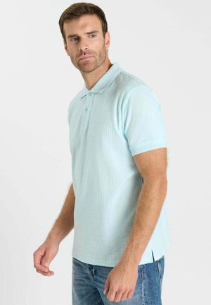 Man met kort bruin haar, gekleed in een lichtblauw gestructureerd poloshirt en een blauwe spijkerbroek, staand en zijwaarts kijkend.