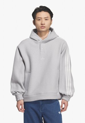 Giovane uomo che indossa una felpa con cappuccio grigio chiaro con tre strisce bianche sulle maniche e pantaloni Adidas blu su sfondo bianco.