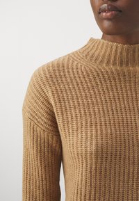 Strickpullover in Hellbraun mit gerippter Textur und hohem Kragen. Nahaufnahme des Schulter- und Brustbereichs.