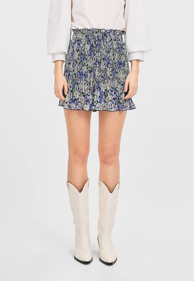 Femme portant un chemisier blanc, une mini-jupe bleue à fleurs et des bottes blanches hauteur genoux, debout devant un fond uni.