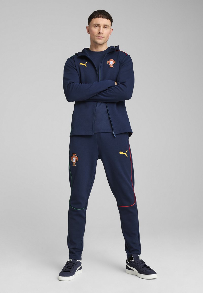 Tamsiai mėlynas treniruočių kostiumas su užtrauktuku ir kelnių, kuriame yra auksinis Puma logotipas, Portugalijos herbas ir žali-raudoni akcentai šonuose.