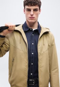 Νέος άνδρας που φοράει μπεζ ζακέτα με φερμουάρ πάνω από σκούρο πουκάμισο τζιν, κρατώντας μαύρο λουράκι στον δεξιό ώμο, κοιτάζοντας εμπρός με ουδέτερη έκφραση.