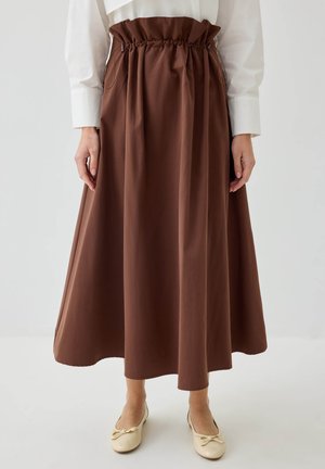 Femme portant une jupe midi marron taille haute avec une taille froncée et des ballerines crème, debout devant un fond clair uni.