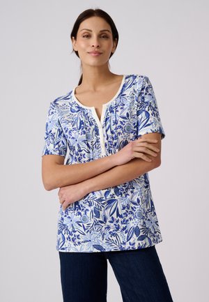 Frau mit dunklen Haaren, die eine weiße kurzärmelige Bluse mit blauen Blumenmustern und dunkle Hose trägt, steht mit verschränkten Armen.