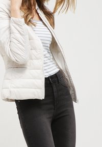 Kvinna som bär en vit quiltad jacka över en vit och blå randig skjorta med svarta jeans med hög midja mot en enfärgad bakgrund.