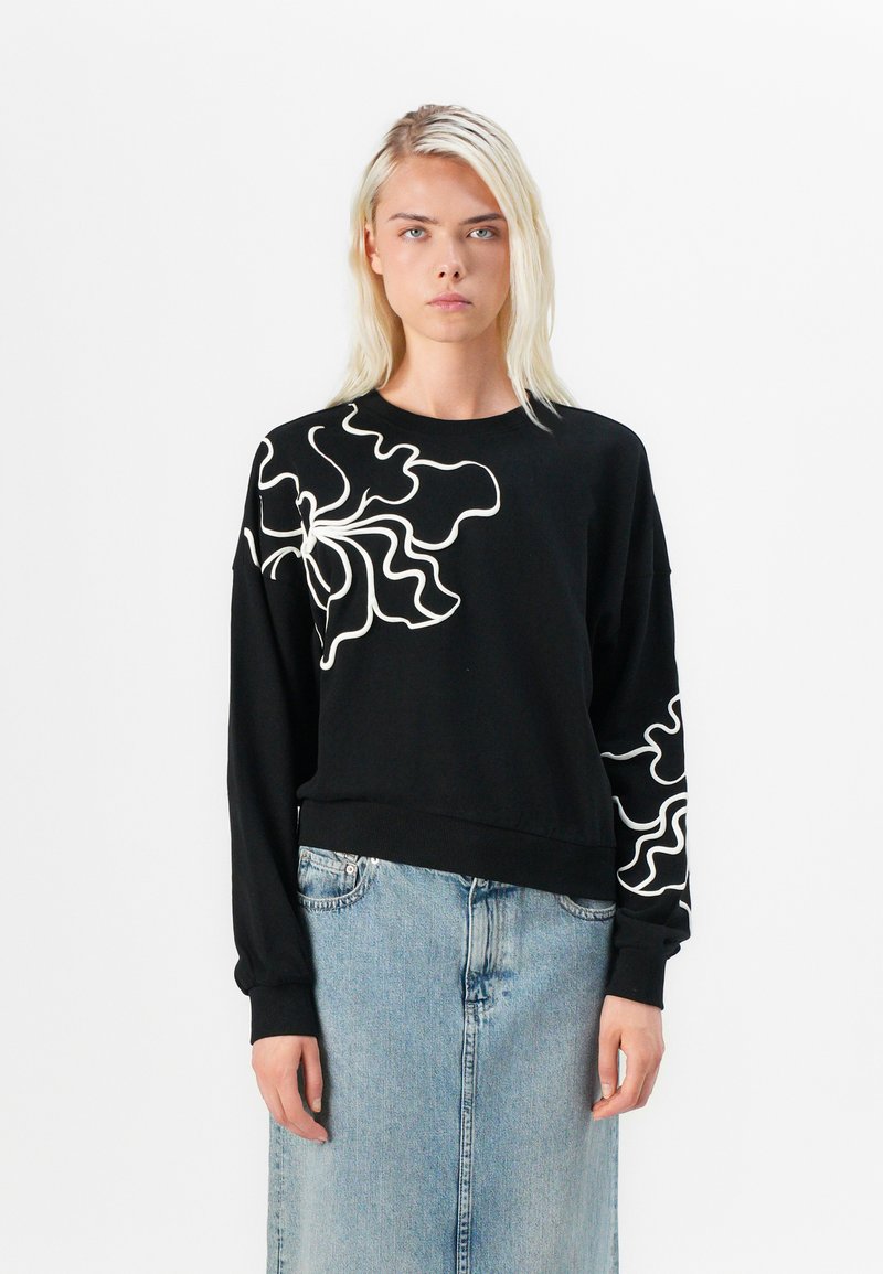Felpa nera con maniche lunghe, caratterizzata da motivi astratti bianchi sulle spalle. Orlo e polsini a costine; indossata con una gonna di jeans.