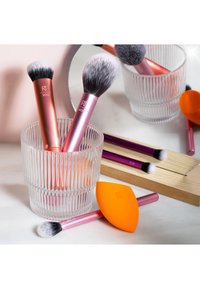Deux pinceaux à maquillage à manche rose dans un verre cannelé, un pinceau à manche violet et une éponge à maquillage orange sur une surface blanche.