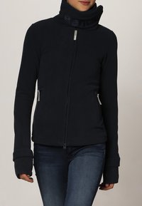 Chaqueta de forro polar de color azul oscuro con cuello alto, cremallera frontal, bolsillos laterales y orificios para los pulgares en los puños para mayor comodidad y calor.