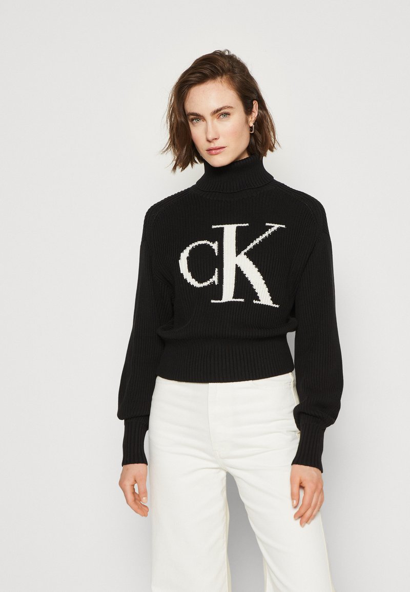 Črn pleten turtleneck pulover z rebrasto teksturo in prevelikim krojem, s poudarjeno velikim belim logotipom "CK" spredaj. V kombinaciji z belimi hlačami.