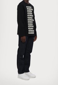 Una maglietta nera a maniche lunghe presenta del testo bianco audace sulla parte anteriore, abbinata a jeans di denim scuri e sneakers bianche, creando un look casual.