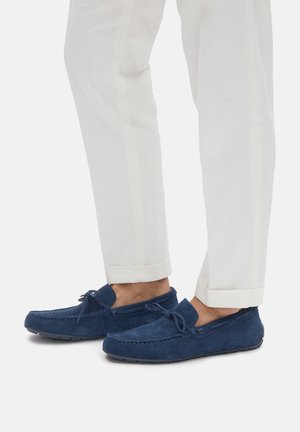 Marineblaue Veloursleder-Slipper mit gesteppten Details und Schnürungsakzenten. Runde Zehenpartie und Gummisohle, getragen zu weißen, aufgekrempelten Hosen.