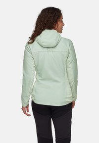 Femme aux cheveux bouclés portant une veste à capuche verte claire et un pantalon noir, vue de dos, sur fond blanc.