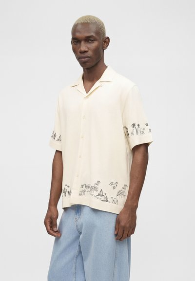 Άνδρας με κοντά ξανθά μαλλιά που φοράει ένα κρεμ πουκάμισο με κοντά μανίκια, με μαύρα σκίτσα ερήμου και ιστιοπλοϊκού, συνδυασμένο με ανοιχτό μπλε τζιν.