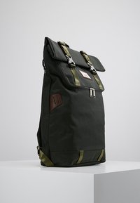 Zielony plecak materiałowy z zamknięciem typu roll-top, regulowanymi paskami, brązowymi skórzanymi akcentami, podwójnymi zamkami błyskawicznymi oraz minimalistycznym designem.