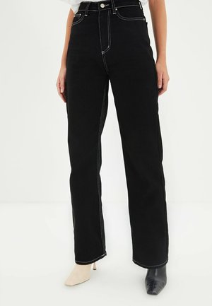 Persoon die zwarte high-waist jeans draagt met witte contrasterende stiksels, gecombineerd met niet-matching beige en zwarte puntige haklaarzen.