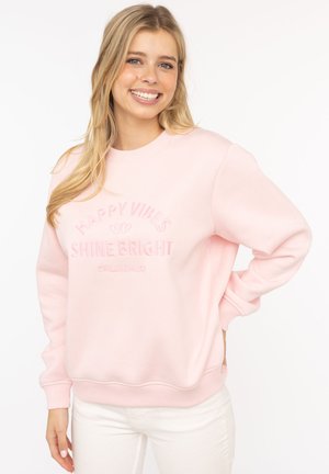 Hellrosa Sweatshirt mit gerippten Bündchen und Saum. Auf der Vorderseite mit erhabenem Text "HAPPY VIBES" und "SHINE BRIGHT" versehen. Weiche Textur.