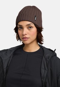 Bruine geribbelde breimuts met een opgerolde rand en een merklabel. Draagt het samen met een zwarte zip-up jasje over een zwart top.