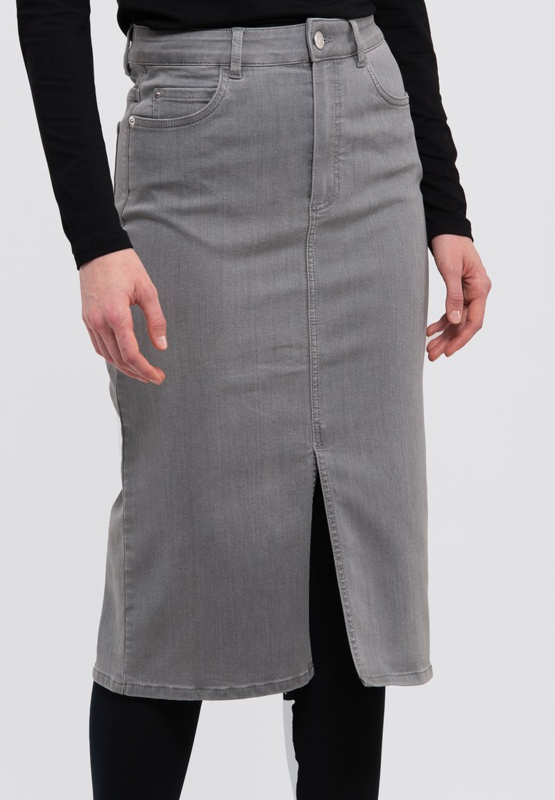 Falda lápiz de mezclilla gris con una abertura frontal, diseño de cinco bolsillos, trabillas para cinturón y costuras en contraste, llevada sobre leggings negros.