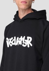 Uomo che indossa un hoodie nero con testo in stile graffiti bianco "DISCLAIMER" e orecchini a cerchio dorati, su uno sfondo chiaro e semplice.