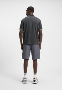 Vyras, dėvintis tamsiai pilką tekstūruotą polo marškinėlį, pilkus kelio ilgio šortus, baltas kojines ir baltus sportinius batelius, stovintis nugara į akis prieš paprastą foną.