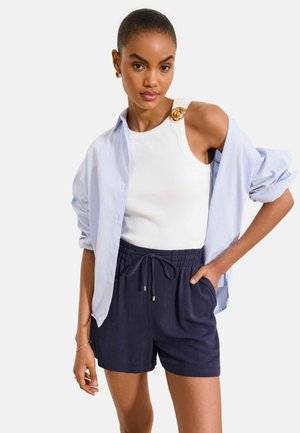 Vrouw draagt een wit mouwloos topje met gouden broche, lichtblauw open hemd en marineblauwe short met trekkoord, poseert met één hand in de zak.