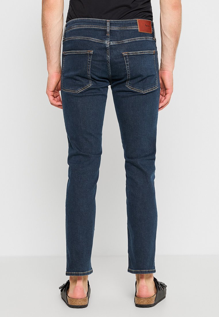 Schmale, dunkelblaue Jeans aus texturiertem Denim-Material, mit fünf Taschen und einem braunen Lederpatch am hinteren Bund.