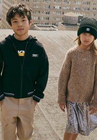 Hoodie zippé noir avec des accents blancs, T-shirt noir, pantalon cargo beige ; pull en laine beige, jupe en sequins argentés et bonnet noir.