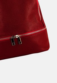 Sac en cuir rouge avec une surface texturée, doté de fermetures éclair argentées et d'une tirette métallique, mis en valeur par des détails de surpiqûres contrastées.