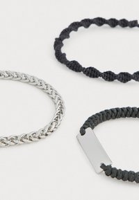 Trīs rokassprādzes: sudraba metāla ķēde, melns pītā aukla un melna aukla ar taisnstūra metāla akcentu. Atšķirīgas tekstūras un krāsas.