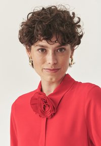 Blusa roja con cuello y un acento de flor tridimensional en el escote. Tela suave con un diseño aerodinámico.