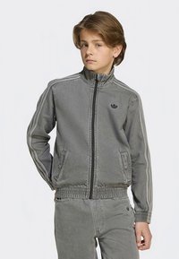 Šedá džínová bunda s vysokým límcem, zapínáním na zip, jemným logem a kontrastními proužky po stranách. Má elastické manžety a pásek v pase.