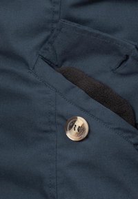 Tessuto blu navy con una texture liscia, caratterizzato da una tasca a patta e un bottone trasparente con due fori. Interno foderato in morbido materiale nero.