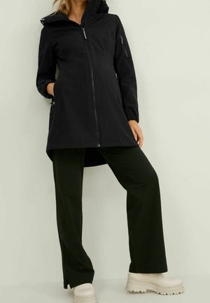 Giacca lunga nera con zip frontale, cappuccio e polsini elasticizzati. Presenta tasche laterali e un taglio dritto. Abbinata a pantaloni neri a gamba larga e stivali beige massicci.