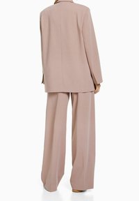 Ljust rosa skräddarsydd kostym med en lång blazer och vida byxor. Slät tygkvalitet med avslappnad passform och minimalistisk design, inga synliga dekorationer.