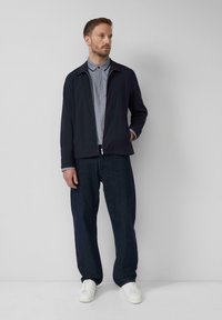 Ein Mann steht an einer schlichten Wand und trägt eine marineblaue Jacke über einem grauen Poloshirt, dunkelblaue Jeans und weiße Sneakers, mit einer Hand in der Tasche.
