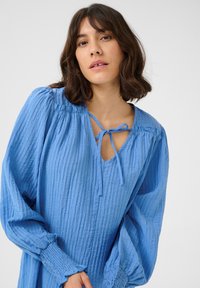 Blouse bleue froissée avec un décolleté en V, de longues manches bouffantes et un détail noué au cou. Texture douce et design fluide.