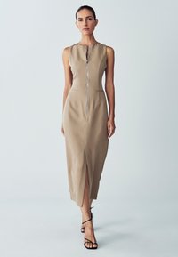 Vestido midi sin mangas beige con cremallera frontal, bolsillos laterales y una abertura delantera. Textura suave con una silueta ajustada.