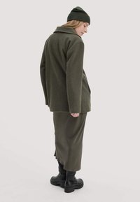 Olivgrüner Wollmantel mit Kragen, kombiniert mit einem passenden olivgrünen Rock und schwarzen klobigen Stiefeln. Glatte Textur, strukturierte Passform und keine sichtbaren Muster.