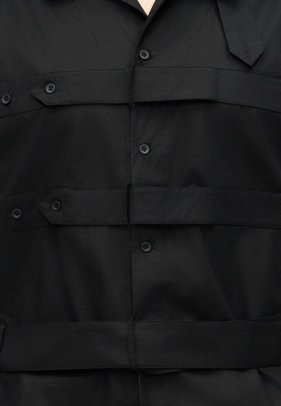 Κοντινό πλάνο μαύρου πουκαμίσου με οριζόντιες λουρίδες με κουμπιά στο στήθος και τη μέση, με μαύρα κουμπιά και γιακά.