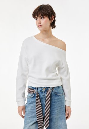 Jonge vrouw met kort bruin haar die een witte off-shoulder trui draagt en spijkerbroek met een gestreepte stoffen riem.
