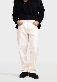 Pantalones blancos de corte holgado con un diseño de pierna recta y estilo de cinco bolsillos, combinados con una camisa negra de botones y zapatos negros.