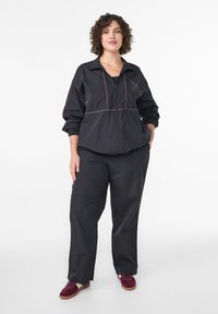 Sieviete ar cirtainiem matiem stāv, ģērbusies melnā džemperī ar rāvējslēdzēju un biksēs, kājās valkā burgundijas krāsas sporta apavus, uz baltas vienkāršas fona.