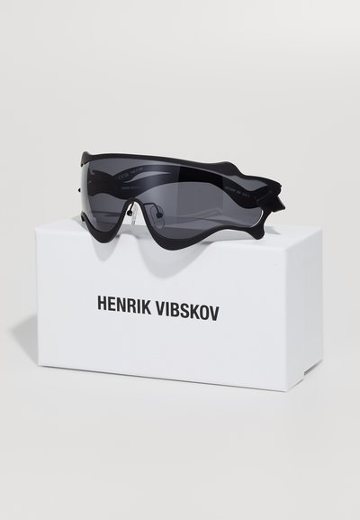 Lunettes de soleil noires surdimensionnées avec un design élégant et des verres teintés, exposées sur une boîte blanche étiquetée "HENRIK VIBSKOV."