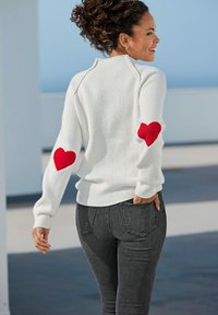 Witte geribde trui met rode hartpatches op de ellebogen, gecombineerd met donkergrijze slim-fit jeans. Het ontwerp heeft een hoge kraag en een gestructureerde stof.