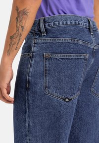 Jeans de mezclilla de cintura alta, con un color azul deslavado, dos bolsillos traseros y una etiqueta de marca con forma de ola en el bolsillo izquierdo.