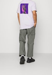 Muž v bledě fialovém tričku s potiskem kaktusu v purpurové a růžové na zádech, šedých cargo kalhotách, bílých ponožkách a černých teniskách.