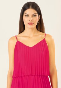 Fuchsia geplisseerde jurk met een V-hals, dunne bandjes met gouden hardware en gelaagd ontwerp. Glad textuur op een lichte achtergrond.