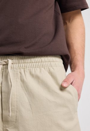 Persoon draagt beige broek met trekkoord, hand in zak en een bruin kort mouwen shirt, lichtjes in de taille gestopt.