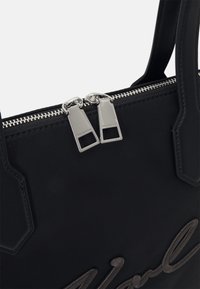 Mustat nailonitote-laukku, jossa on hopeiset kaksinkertaiset vetoketjut, brodeerattu logo ja rakenteelliset kahvat. Silkinpehmeä pinta minimalistisella suunnittelulla.